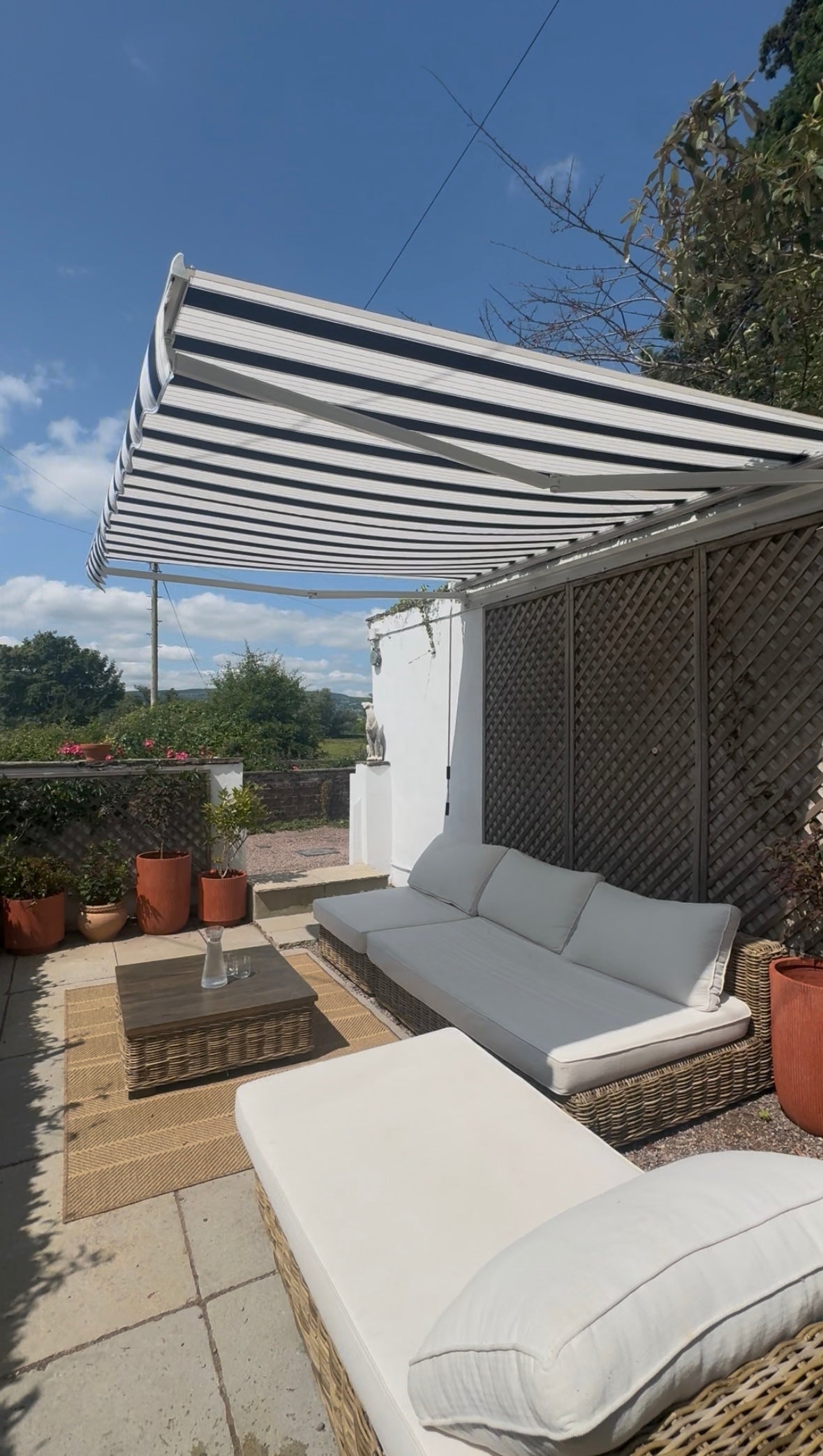 Outdoor patio with wicker furniture, striped awning, and potted plants under a clear blue sky.